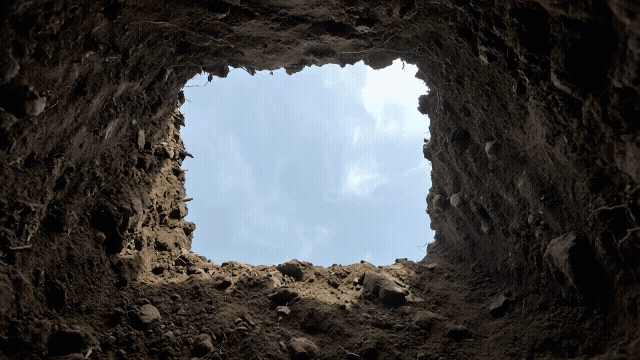 Gen-4_5 A rectangular, hand-dug earthen pit extends downwards, its rough, dark brown soil walls textured with small stones, root fragments, and uneven surfaces, leading to a square opening of bright s.mp4.gif