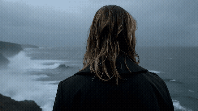 Gen-4_5 over the shoulder of a woman with wet blonde hair facing the ocean the camera cranes back and up to reveal she's standing at the edge of a cliff, stormy waves crashing in the distance below h.mp4.gif